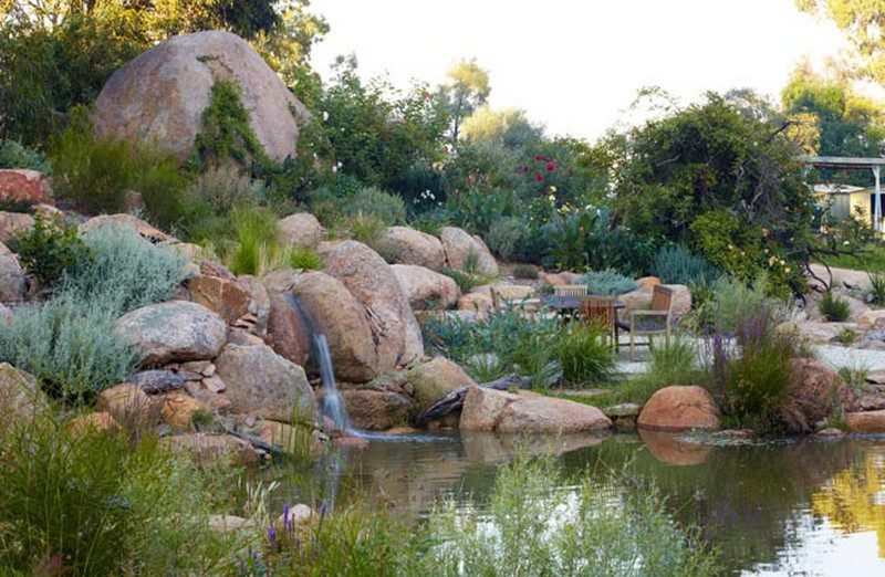 Natural swimming pond landscaped with large boulders, waterfall feature, and garden seating in a lush backyard setting.